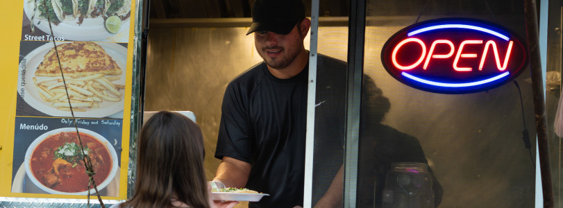 Food truck worker handing dish to a customer near an open neon sign